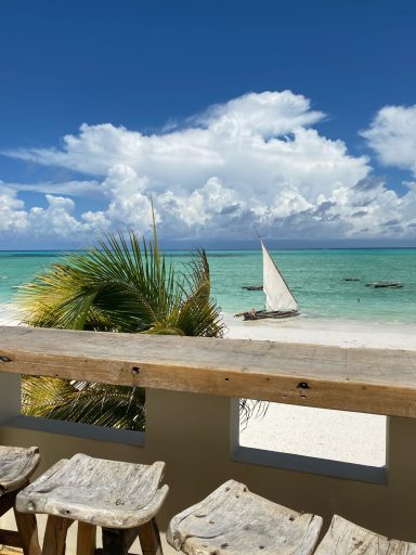 Strandblick mit einem Segelboot und Palmen unter blauem Himmel und weißen Wolken.