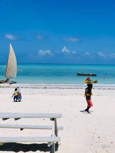 Zwei Personen am Strand mit Booten und klar blauem Wasser im Hintergrund.