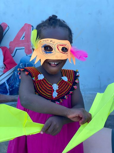Ein lächelndes Kind trägt eine bunte Maske und hält leuchtend gelbe Papierblätter.