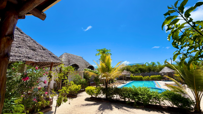 Tropischer Garten mit Pool und palmenbestandenen Hütten unter blauem Himmel.